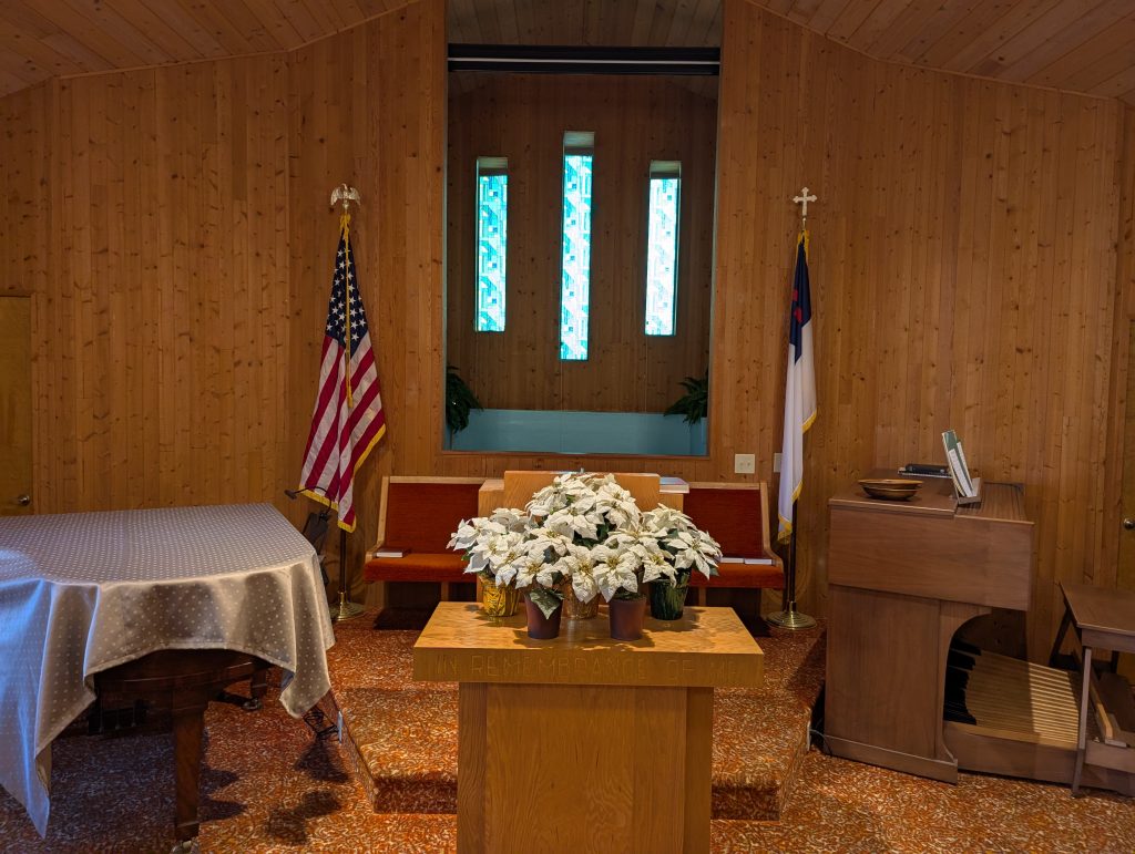 Church interior with wooden walls, stained glass windows, flags behind pews, and an altar featuring white flowers. A draped piano and an organ are on either side.