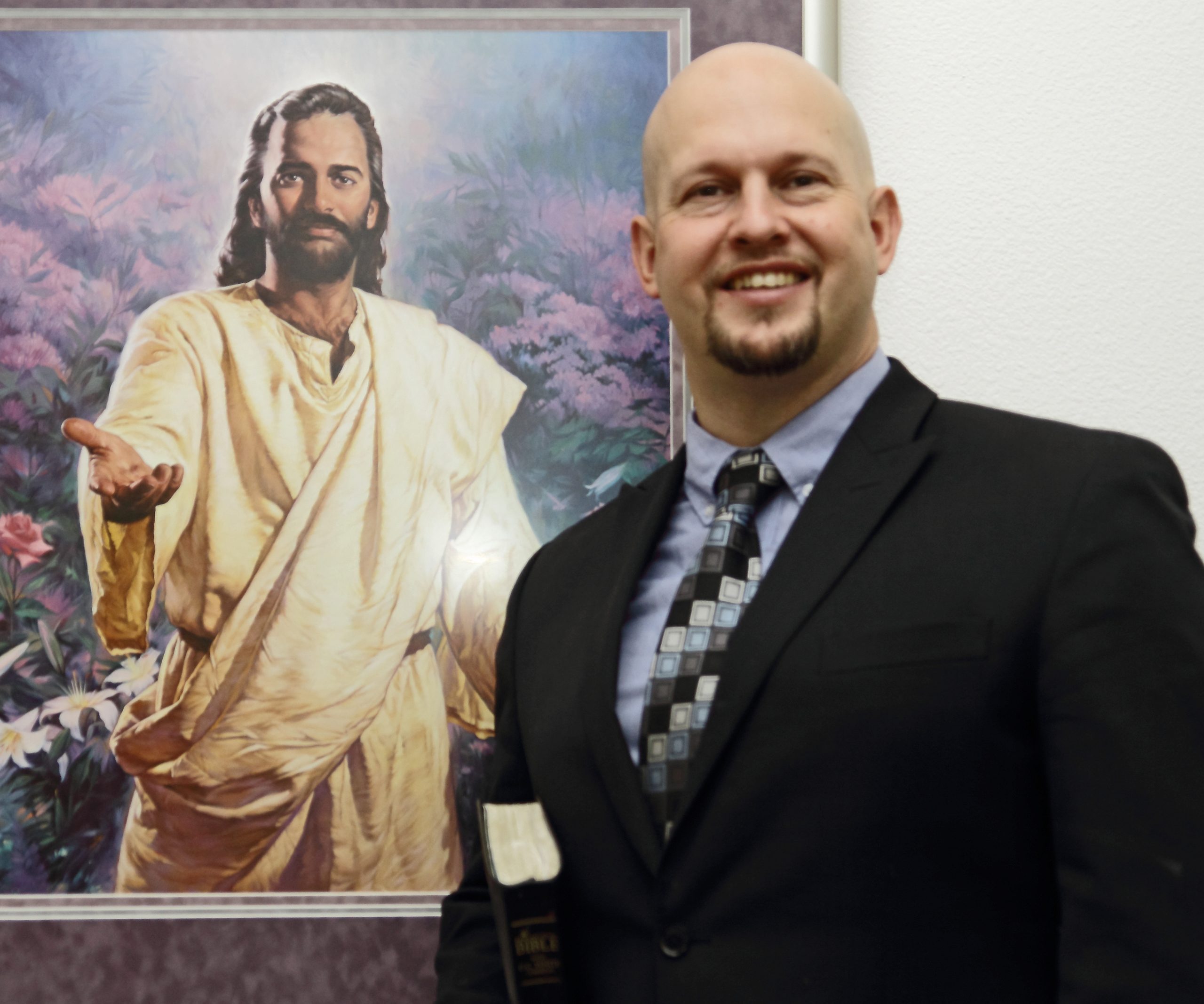 A man in a suit holding a Bible stands and smiles in front of a framed painting of Jesus with an outstretched hand.